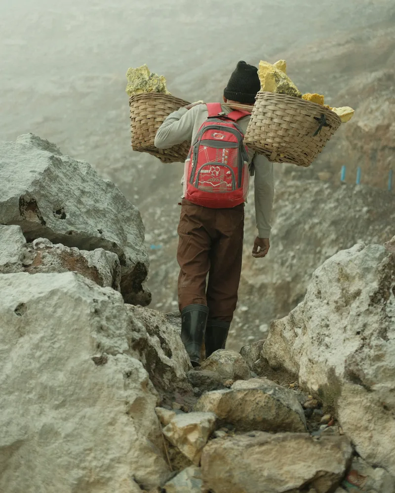Sulfur miner in boots climbing the rocky crater path with two baskets